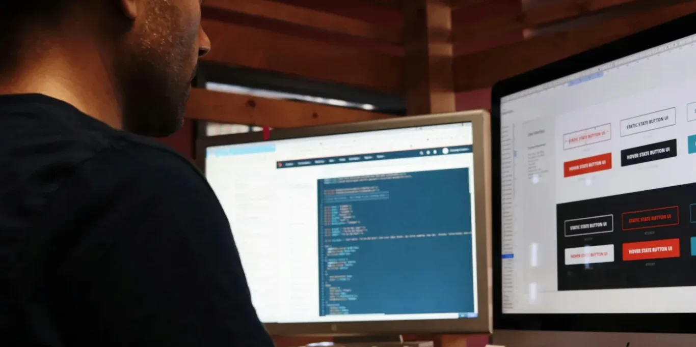Individuelle Programmierung Junger Mann sitzt vor 2 Bildschirmen und liest Computercode